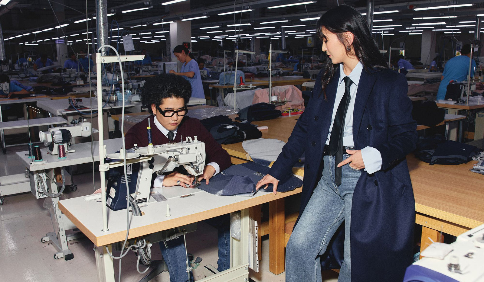 Person using a sewing machine in a factory setting with another person observing.