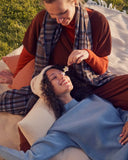 Smiling women relax on a blanket outdoors, one in a plaid scarf, the other in a rib-knit and beanie