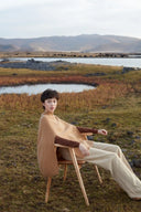 Model in camel ribbed cashmere poncho from traceable cashmere brands, seated outdoors by a lake, side view
