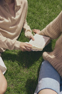 Two women sitting on grass in lightweight cashmere for summer, beige polo knit and tan ribbed sweater, reading a book