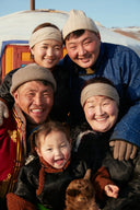 Smiling Mongolian herder family in traditional deels and beige knit hats outside a ger, showing where cashmere is from