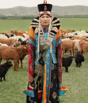 Woman in traditional Mongolian deel and headdress standing among a goat herd on green steppe, things to do in Mongolia