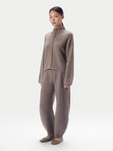 A model wearing a matching taupe knit two-piece set consisting of a mock-neck long-sleeve top with a braided front detail and wide-leg trousers, paired with brown suede flats, against a light gray studio background.