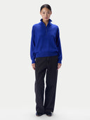 A model stands against a light gray studio background wearing a bright royal blue quarter-zip mock-neck sweater and dark charcoal wide-leg trousers.