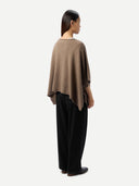 A woman shown from the back wearing a taupe-brown draped poncho-style top and loose black wide-leg trousers, standing against a plain light gray studio background.