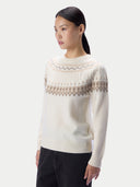 A woman modeling a cream-colored knit sweater with a brown Fair Isle pattern across the chest and shoulders, paired with black pants, against a light gray studio background.