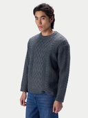 A man wearing a dark gray cable-knit crewneck sweater and blue jeans, standing against a light gray studio background.