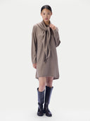 A model wearing a taupe knit wrap dress with a large bow detail at the neckline, paired with black knee-high boots, against a plain light gray background.