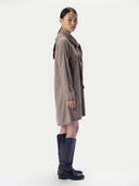 A model shown in profile wearing a taupe-brown wool coat with a shawl collar, paired with dark navy knee-high boots, against a plain light gray studio background.