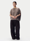 A model stands against a light gray studio background wearing a loose taupe knit top with wide draped sleeves and dark wide-leg trousers.