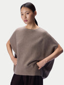 A woman modeling a taupe-brown knit sweater with a wide, draped batwing silhouette and a crew neckline, paired with dark pleated trousers, against a light gray studio background.