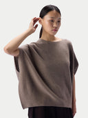 A woman modeling a loose-fitting, short-sleeve knit top in taupe brown against a light gray studio background.