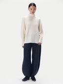 A model stands against a plain light gray studio background wearing a cream turtleneck sweater, dark navy wide-leg knit pants, and dark loafers.