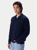 A man wearing a dark navy blue zip-up knit polo jacket over a white t-shirt and light blue jeans, photographed against a solid light gray background.