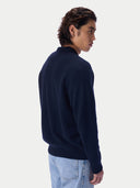 A man wearing a dark navy blue knit sweater and light blue jeans, shown from a side-back profile against a clean off-white studio background.