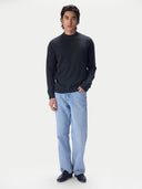 A man modeling a dark charcoal gray mock neck knit sweater, paired with light blue jeans and dark loafers, standing against a solid off-white studio background.