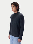 A man wearing a dark charcoal gray knit sweater and light blue jeans, shown in a side-profile studio shot against a light gray background.