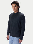 A man wearing a dark charcoal gray knit sweater and light blue jeans, standing against a light gray studio background.