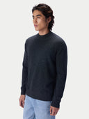 A man wearing a dark charcoal gray knit sweater and light blue jeans, standing against a light gray studio background.