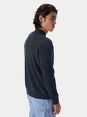 A man wearing a dark charcoal gray knit mock neck sweater and light blue jeans, shown from a side-back profile against a light gray studio background.