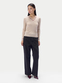 A woman modeling a beige ribbed V-neck sweater with thin red trim, paired with dark charcoal wide-leg trousers and burgundy pointed heels, against a plain light gray studio background.