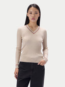 A woman modeling a ribbed beige V-neck sweater with thin red and green trim, paired with black pants, against a light gray studio background.