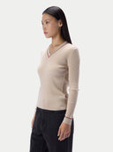A woman wearing a ribbed beige V-neck sweater with thin red stripe trim on the collar and cuffs, paired with black pants, photographed against a light gray studio background.