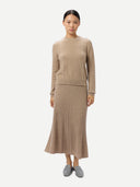 A model stands against a light gray studio background wearing a beige knit sweater and matching pleated midi skirt, paired with gray slip-on shoes.