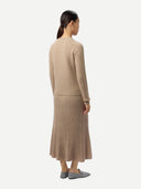 back view of a woman wearing a beige sweater and pleated skirt on a white background.