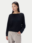 A woman modeling a black long-sleeve knit sweater with a wide boat neckline, paired with light beige trousers, against a solid light gray background.