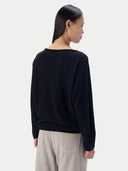 A woman shown from the back wearing a black long-sleeve knit top and light beige trousers against a plain light gray studio background.