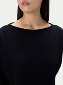 Close-up of a woman wearing a solid black long-sleeve top with a wide boat neckline, shown against a plain light gray background.