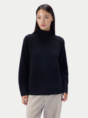 A woman wearing a black loose-fit turtleneck knit sweater with raglan sleeves and light gray wide-leg trousers, standing against a light gray studio background.