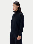 A woman in a black long-sleeve quarter-zip mock neck sweater and matching black trousers, shown in profile against a light gray studio background.