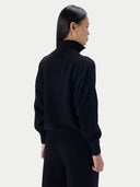 A model shown from the back wearing a black mock neck knit sweater and matching black trousers against a light gray studio background.