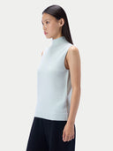 A woman shown in side profile wearing a sleeveless mock-neck knit sweater in a very light icy blue, paired with dark pants, against a plain light gray studio background.