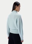 A woman shown from the back wearing a light mint green ribbed knit mock-neck sweater and black pants, photographed against a clean off-white studio background.