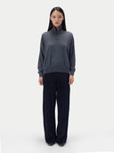 A model stands against a light gray studio background wearing a dark gray quarter-zip mock-neck sweater and dark navy wide-leg trousers.
