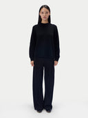 A model stands against a light gray studio background wearing a black mock-neck knit sweater and matching black wide-leg trousers.