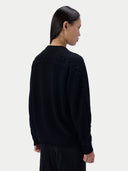 A woman shown from the back wearing a black knit sweater with textured cable detailing across the shoulders and down the sleeves, set against a plain light gray studio background.