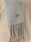 A close-up shot of the fringed end of a light gray cashmere scarf featuring a dark blue cursive monogram "OF" embroidered near the edge, draped over a beige garment. #color_default