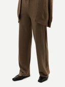 A close-up of a person wearing brown wide-leg trousers with a matching textured brown cardigan or jacket draped over one shoulder, paired with black pointed flats, against a light gray studio background.