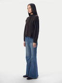 A woman stands in profile wearing a dark brown mock-neck button-up cardigan, blue wide-leg jeans, and black shoes against a plain light gray studio background.