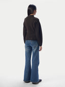A model viewed from the back wearing a dark brown mock-neck knit sweater and blue wide-leg jeans, standing against a plain light gray studio background.