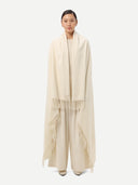 front view of a woman standing wrapping a white cashmere blanket with fringes over a white outfit on a light gray background.