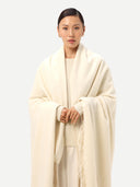 close up front view of a woman standing wrapping a white cashmere blanket with fringes over a white outfit on a light gray background.