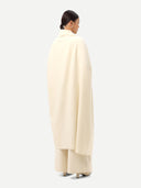 back view of a woman standing wrapping a white cashmere blanket with fringes over a white outfit on a light gray background.