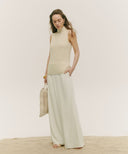 Woman wearing a beige sleeveless top and light green pants, holding a woven bag on a plain background