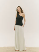 A full-length front view of a person with long, wind-swept brown hair standing on a sandy surface against a cream background. They are wearing a black one-shoulder knit top tucked into flowing, cream-colored wide-leg trousers. #color_black