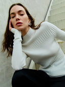 A woman sitting with her hands behind her head, wearing a high-neck ribbed cotton silk cashmere blend sweater in a light color. #color_whisper-white